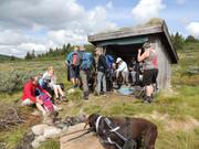 Bildet viser gruppen samlet for lunsj ved og i en gapahuk bygd av treverk. I forgrunnen står hunden Taro, som var med på turen